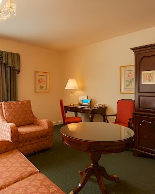 A cozy hotel suite with a round wooden table, pink patterned sofa and chair, a dark wooden armoire, desk with laptop, lamp, framed art, and a chandelier, all near a window with draped curtains.