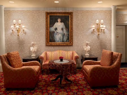 A luxurious hotel lobby sitting area with four dusty rose chairs, a small central table, ornate wall art, and vintage-style lamps.