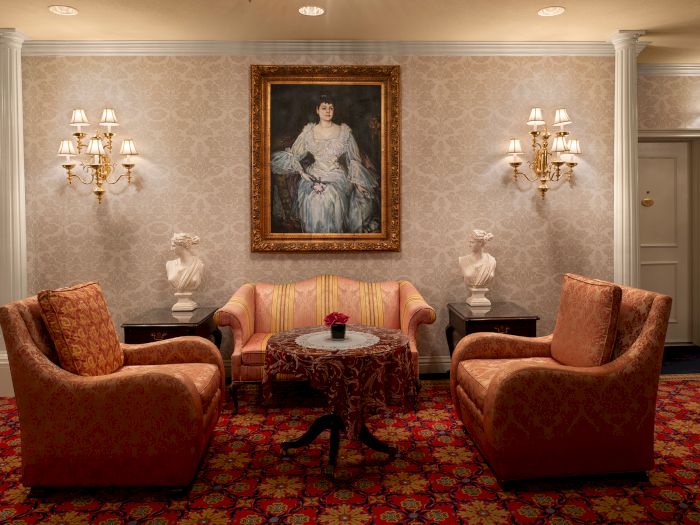 A luxurious hotel lobby sitting area with four dusty rose chairs, a small central table, ornate wall art, and vintage-style lamps.