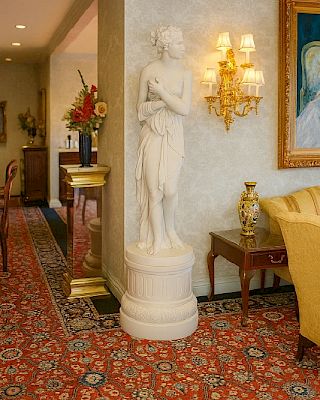 A classical marble statue of a draped woman stands on a pedestal in a luxurious hallway, with ornate lamps, framed art, and a red patterned carpet.