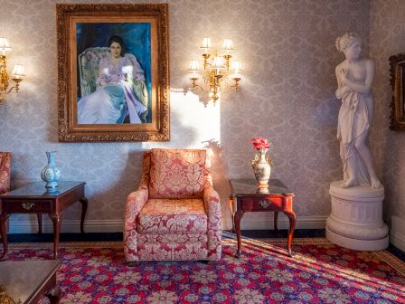 A richly decorated living room with ornate wallpaper, velvet patterned chairs, a framed portrait, statues, lamps, and a red patterned rug.