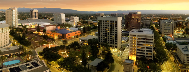 A wide aerial view of a sunny cityscape with mid-rise buildings, trees, streets, and a plaza, bathed in warm sunset light.