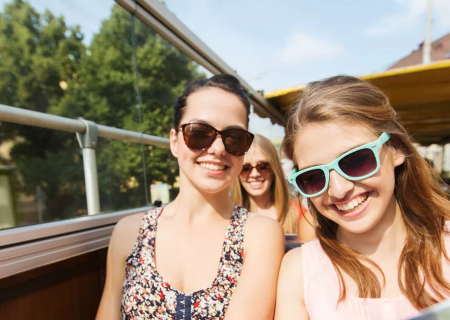 Three smiling friends ride a sunny open-top bus, wearing sunglasses, enjoying a day out.