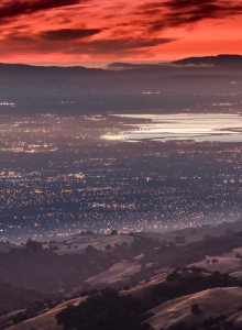 A city glows in a valley below a dramatic red sunrise, with rolling hills in the foreground and distant mountains beneath a fiery sky.
