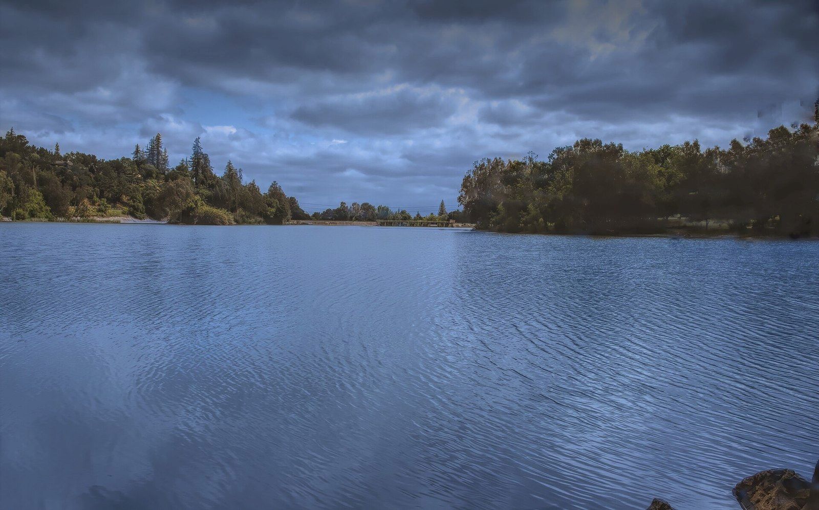 A calm lake under a cloudy sky, framed by dense green trees and conifers along the shore.