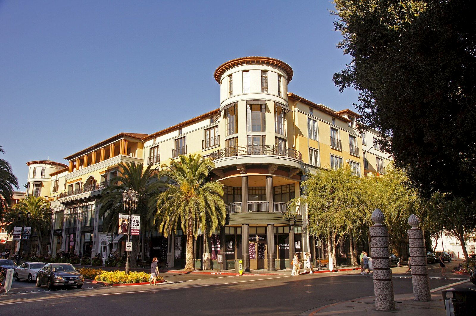 A sunny street with a grand yellow hotel building, curved balconies, palm trees, and pedestrians walking by.