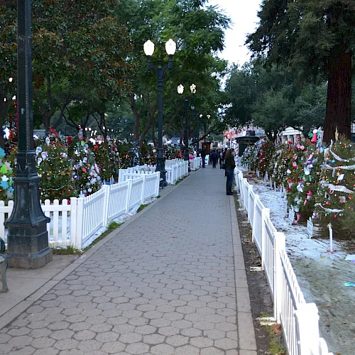 A park walkway is lined with white fences, holiday trees, and colorful lights and decorations during a festive fair.