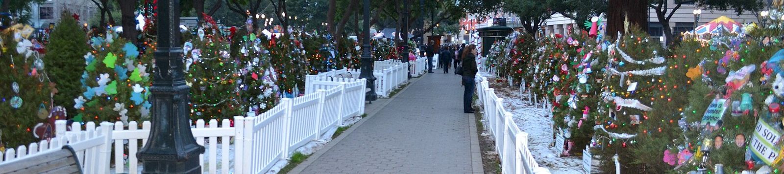 A festive outdoor walkway lined with white fences, lit trees and colorful decorations, with people walking through at dusk.