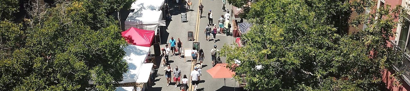 A sunny campus walkway packed with people, vendor tents, and umbrellas between leafy buildings with mountains in the distance.